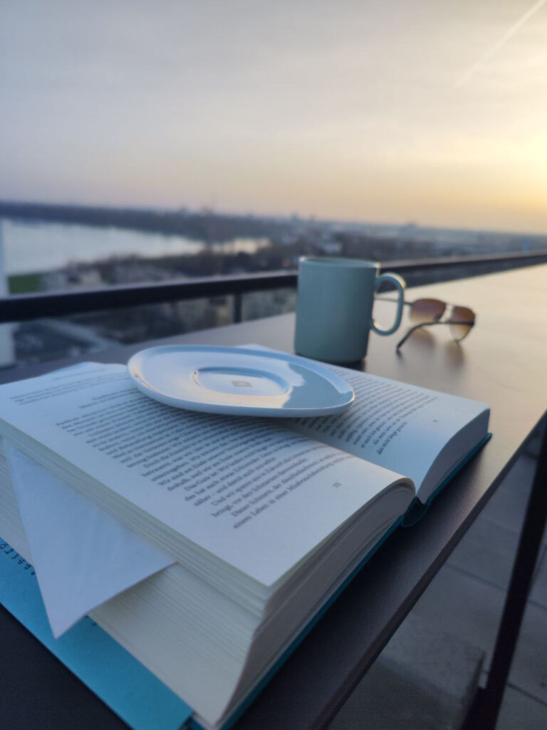 Eine Nahaufnahme eines geöffneten Buches auf einer Balkonbrüstung mit Blick auf ein Gewässer und eine Stadt im Hintergrund bei Sonnenuntergang. Auf dem Buch liegt eine Untertasse mit einem goldenen Muster. Neben dem Buch steht eine hellblaue Tasse und eine Sonnenbrille. Der Himmel ist in orangefarbenen und blauen Tönen gehalten.