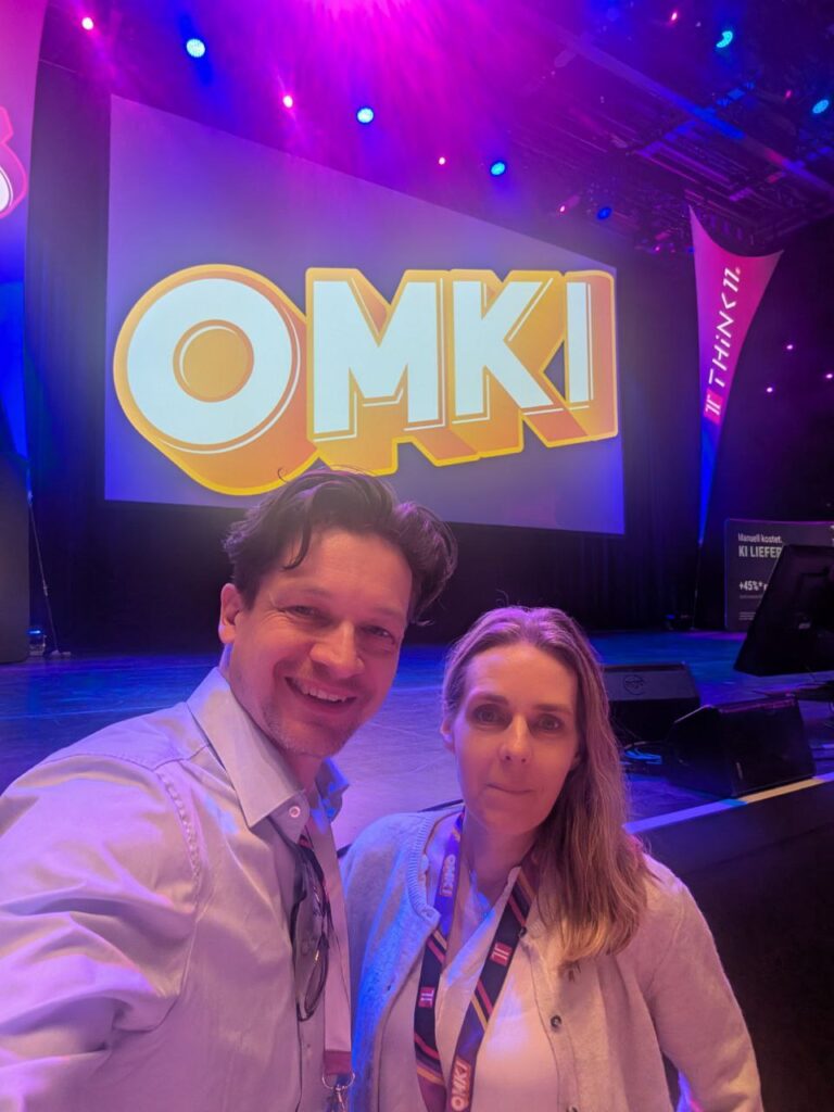 Ein Selfie-Foto von einem Mann und einer Frau, die vor einer großen Leinwand stehen, auf der der Schriftzug "OMKI" in gelben Buchstaben auf blauem Hintergrund zu sehen ist. Die Frau trägt einen hellen Pullover und einen lila-orange gestreiften Schal, der Mann ein weißes Hemd. Der Mann lacht und blickt in die Kamera, während die Frau einen ernsteren Ausdruck hat. Der Hintergrund ist ein Konzertsaal mit lila Beleuchtung.