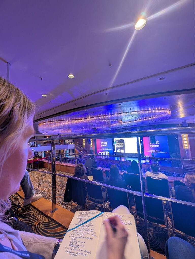 Eine Ansicht von oben auf eine Frau, die in einem Konzertsaal sitzt und Notizen macht. Die Frau hat blonde Haare und trägt ein lila-orange gestreiftes Band. Im Hintergrund ist eine große Bühne mit einer Leinwand zu sehen, auf der der Schriftzug "OMKI SUMMIT" in weißen Buchstaben auf blauem Hintergrund steht. Der Saal ist mit lila und blauer Beleuchtung beleuchtet.