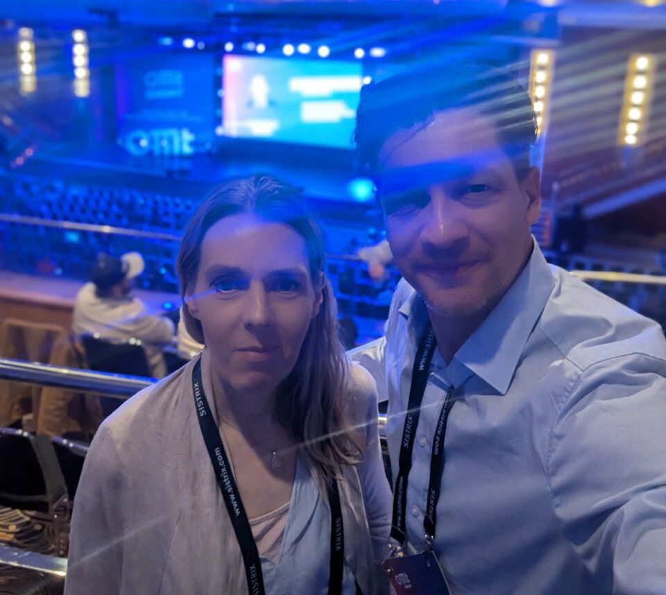 Ein Selfie-Foto von Federico Zimmermann und Christine Neal-Eßmann, die vor einer großen Leinwand stehen, auf der der Schriftzug "OMT SUMMIT" in weißen Buchstaben auf blauem Hintergrund zu sehen ist. Die Frau trägt einen hellen Pullover und einen lila-orange gestreiften Schal, der Mann ein weißes Hemd. Der Mann lacht und blickt in die Kamera, während die Frau einen ernsteren Ausdruck hat. Der Hintergrund ist ein Konzertsaal mit lila Beleuchtung.